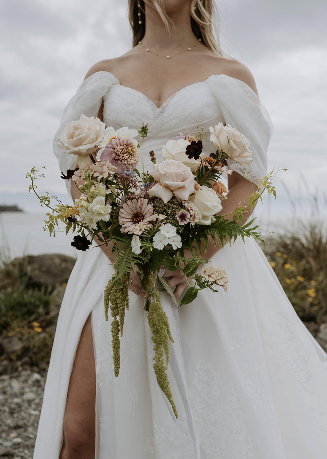 Bride in an off-shoulder white gown holding a bouquet of assorted flowers, standing outdoors with a cloudy sky backdrop.